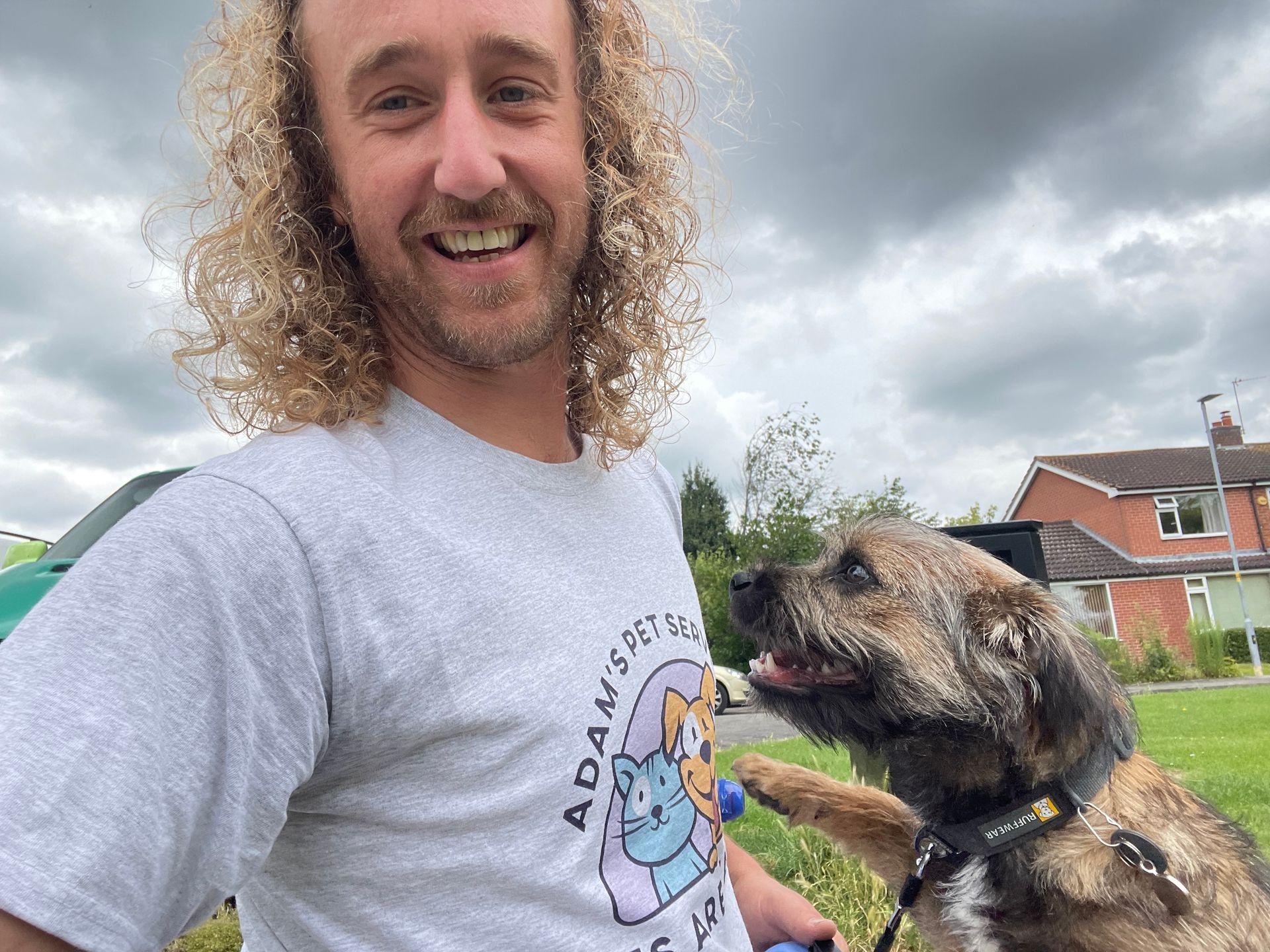 Smiling person with curly hair and a dog outdoors, cloudy sky and houses in the background.